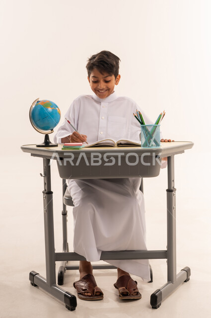 الاجتهاد للوصول إلى النجاح والتفوق، موسم العودة للمدارس، بورتريه لطالب عربى خليجي عماني يرتدي الزي التقليدي يجلس على طاوله مكتبية ، ولد سعودي يقوم بحل الواجبات والمراجعة، قرطاسية مدرسية ومجسم كرة أرضية، خلفية بيضاء