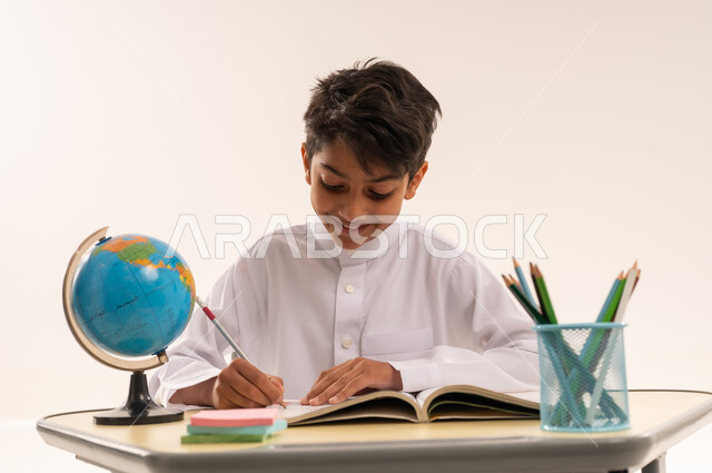 مفهوم العودة إلى المدارس، الاجتهاد للوصول إلى النجاح والتفوق، بورتريه مقرب لطالب عربى خليجي عماني يرتدي الزي التقليدي يجلس على طاوله مكتبية ، ولد سعودي يقوم بحل الواجبات والمراجعة، قرطاسية مدرسية ومجسم كرة أرضية، خلفية بيضاء