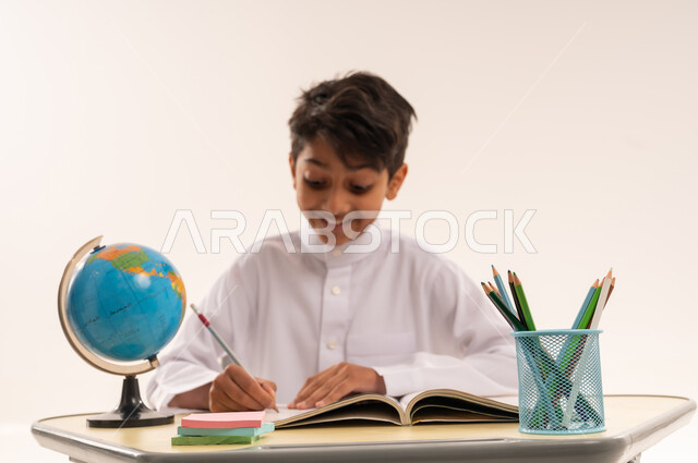 Back to school season, hard work to achieve success and excellence, close-up portrait of an Arab Gulf Omani student wearing traditional dress sitting at an office table, a Saudi boy doing homework and reviewing, school stationery and a globe, white background