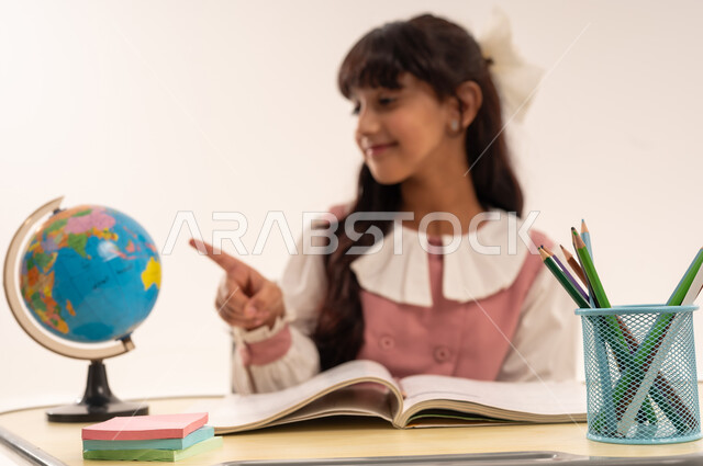 Developing educational curricula in the Sultanate of Oman, the concept of research and exploration around the world, a close-up portrait of an Omani Gulf Arab student wearing a school uniform sitting at a desk pointing at a globe, a Saudi girl studying school lessons, white background