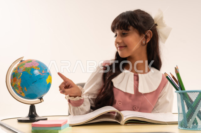 Quality of effective learning methods and the use of tools and aids, the concept of research and exploration around the world, a close-up portrait of an Arab Gulf Omani student wearing a school uniform sitting at an office table pointing at a globe, a Saudi girl studying school lessons, white background