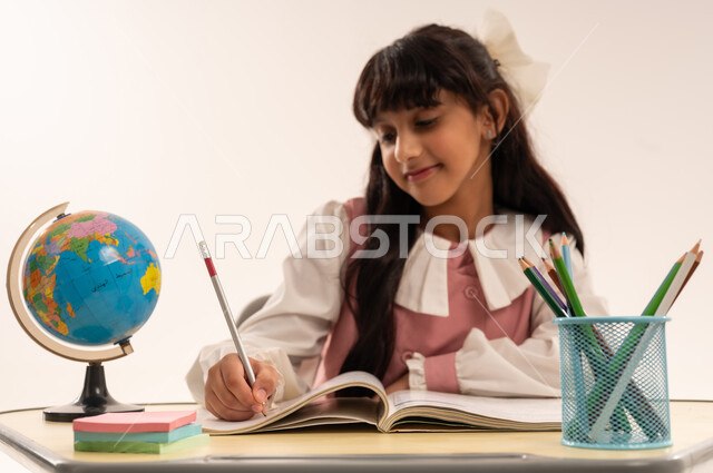 موسم العودة للمدارس، المقررات والمناهج التعليمية الحديثة والمطورة، مفهوم الاجتهاد وطلب التفوق، بورتريه مقرب لطالبة عربية خليجية عمانية ترتدي الزي المدرسي تجلس على طاوله مكتبية تقوم بكتابة شيء ما، بنت سعودية تقوم بمذاكره الدروس المدرسيه، خلفية بيضاء