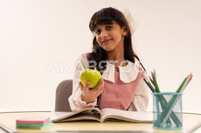 موسم العودة للمدارس، الاجتهاد للوصول إلى النجاح والتفوق، بورتريه مقرب لطالبة عربية خليجية عمانية ترتدي الزي المدرسي تجلس في مقعدها تنظر إلى الكاميرا بإيماءات الاندماج والسعادة، حل الواجبات والمراجعة، بنت سعودية تقوم بامساك تفاحه خضراء، خلفية بيضاء