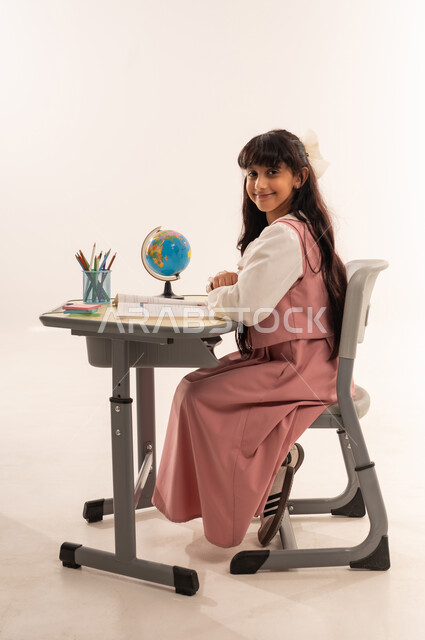 الاجتهاد وطلب العلا والمثابرة والحرص على النجاح، بنت سعودية تنظر اليى الكاميرا بايماءات  السعاده و السرور، بورتريه من الجانب لطالبة عربية خليجية عمانية ترتدي الزي المدرسي تجلس على الطاولة تقوم بالمذاكرة للاختبارات والاستعداد للامتحان، خلفية بيضاء