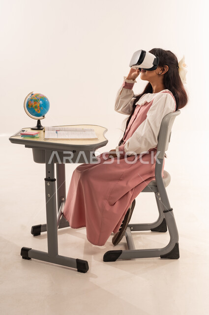 The concept of distance education using modern technology, a Saudi female student sitting at a study table and pointing at something, a side portrait of an Arab Gulf Omani female student wearing a school uniform enjoying watching through virtual reality glasses, white background