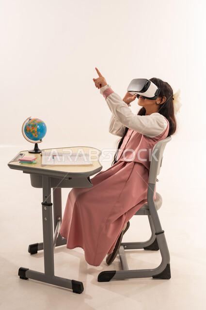 Development of teaching methods using modern advanced technologies, full-length portrait of an Arab Gulf Omani girl wearing a school uniform sitting on a study chair wearing 3D virtual reality glasses looking at something with gestures of integration and concentration, white background