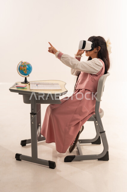 تطور أساليب التعليم باستخدام التقنيات الحديثة المتطورة، طالبة سعودية تجلس على طاولة الدراسة و تشير الى شيء ما، بورتريه من الجانب لتلميذة عربية خليجية عمانية ترتدي الزي المدرسي تستمتع بالمشاهدة عبر نظارة الواقع الافتراضي، خلفية بيضاء