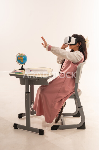 Integrating modern technologies into academic life, a Saudi student sitting at a study table, advanced academies and schools, following new educational methods, a side portrait of an Arab Gulf Omani student wearing a school uniform enjoying watching through virtual reality glasses, white background