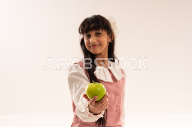 الفطور الصباحي الصحي و المفيد، النظر إلى الكاميرا بإيماءات السعادة و السرور,تلميذة سعودية تستعد للعام الدراسي الجديد, بورتريه مقرب لفتاة عربية خليجية عمانية ترتدي الزي المدرسي تحمل بيدها تفاحة خضراء, الجسم بالطول الكامل, خلفية بيضاء