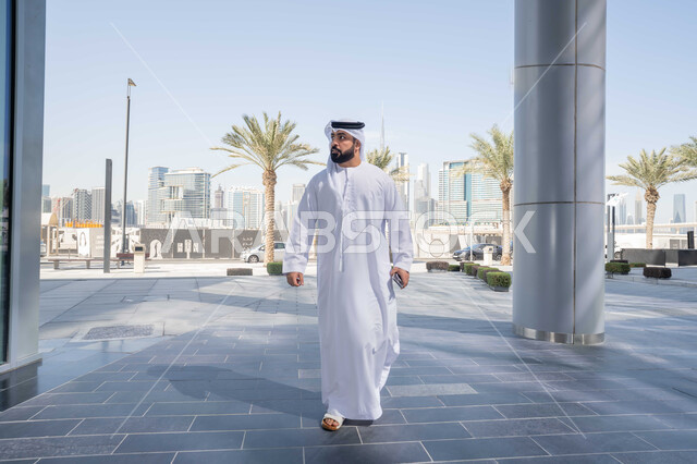 الفخر والاعتزاز بالتقدم المعماري ، النظر بجانباً الى شئ ما ، قضاء وقت ممتع ف الامارات ، الاستمتاع بالاجواء فى الطبيعة ، التجول فى المدينة ، النمو العمراني ، استنشاق الهواء النقي في الخارج, رجل عربي خليجي اماراتي يرتدي الكندورة والغترة البيضاء ينظر الى شيء ما في وقت النهار .