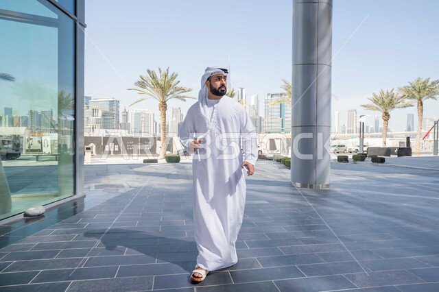 النظر بجانباً الى شئ ما ، قضاء وقت ممتع ف الامارات ، الاستمتاع بالاجواء فى الطبيعة ، التجول فى المدينة ، النمو العمراني ، الفخر والاعتزاز بالتقدم المعماري ،  استنشاق الهواء النقي في الخارج, رجل عربي خليجي اماراتي يرتدي الكندورة والغترة البيضاء ينظر الى شيء ما في وقت النهار .