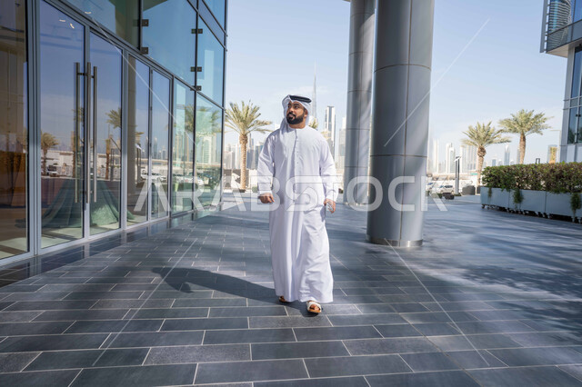 Spending a pleasant time in the Emirates, enjoying the natural atmosphere, strolling around the city, urban growth, pride in architectural progress, and breathing fresh air outside. An Emirati Gulf Arab man wearing a kandura and white ghutra looks at something during the day.