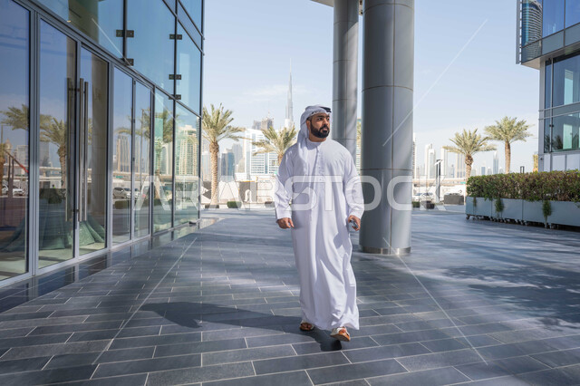 Enjoying nature, strolling around the city, urban growth, having a great time in the Emirates, taking pride in architectural progress, breathing fresh air outside, an Emirati Gulf Arab man wearing a kandura and white ghutra looking at something during the day.