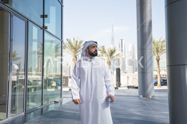 الخروج فى الهواء الطلق ، الفخر والاعتزاز بالتقدم المعماري ، النظر بجانباً الى شئ ما ، قضاء وقت ممتع ف الامارات ، الاستمتاع بالاجواء فى الطبيعة ، النمو العمراني ، استنشاق الهواء النقي في الخارج, رجل عربي خليجي اماراتي يرتدي الكندورة والغترة البيضاء ينظر الى شيء ما في وقت النهار .