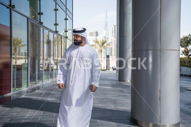 Walking outside, taking pride in architectural progress, looking off to the side at something, having a good time in the Emirates, enjoying the natural atmosphere, urban growth, breathing fresh air outside, an Emirati Gulf Arab man wearing a kandura and white ghutra looking at something during the day.