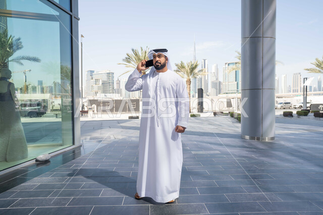 الدردشة مع الاصدقاء ، تصفح المواقع الالكترونية، نزهة وجولة فى الهواء الطلق ، استخدام التكنولوجيا والتقنيات الحديثه، ايماءات السعادة والسرور، رجل عربي خليجي اماراتي يرتدي الكندورة والغترة البيضاء، ينظر بتعابير السرور يقوم باستخدام الهاتف المحمول ، التسوق عبر تطبيقات الاونلاين .