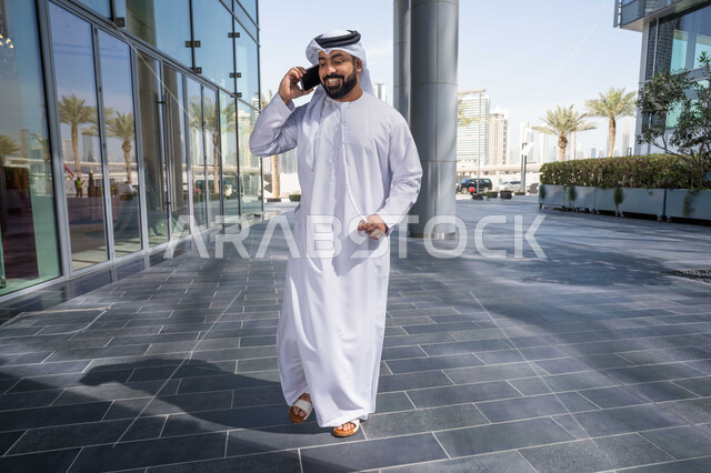 Using modern technology and techniques, chatting with friends, gestures of happiness and pleasure, a walk and a walk in the open air, documenting happy moments, an Emirati Gulf Arab man wearing a white kandura and ghutra, looking with happy expressions, using a mobile phone, shopping via online applications.
