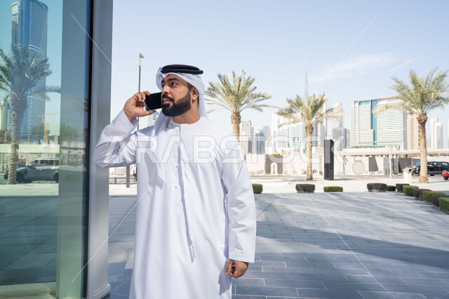 Internet programs and applications, chatting with friends, gestures of happiness and pleasure, using modern technology and techniques, a walk and a tour in the open air, documenting happy moments, an Arab Gulf Emirati man wearing a white kandura and ghutra, looking with expressions of pleasure using a mobile phone, shopping via online applications.