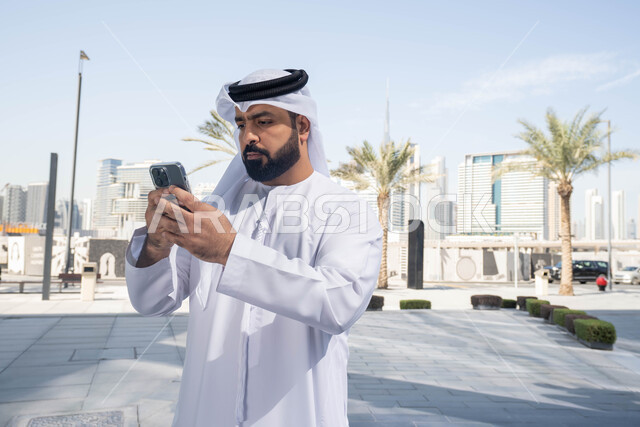 التقاط الصور التذكارية ، ايماءات السعادة والسرور، استخدام التكنولوجيا والتقنيات الحديثه، نزهة وجولة فى الهواء الطلق ، برامج وتطبيقات الانترنت ، الدردشة مع الاصدقاء ، توثيق اللحظات السعيدة، رجل عربي خليجي اماراتي يرتدي الكندورة والغترة البيضاء، ينظر بتعابير السرور يقوم باستخدام الهاتف المحمول ، التسوق عبر تطبيقات الاونلاين .