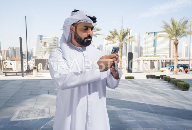 توثيق اللحظات السعيدة، نزهة وجولة فى الهواء الطلق ، استخدام التكنولوجيا والتقنيات الحديثه، الدردشة مع الاصدقاء ، ايماءات السعادة والسرور، رجل عربي خليجي اماراتي يرتدي الكندورة والغترة البيضاء، ينظر بتعابير السرور يقوم باستخدام الهاتف المحمول ، التسوق عبر تطبيقات الاونلاين .