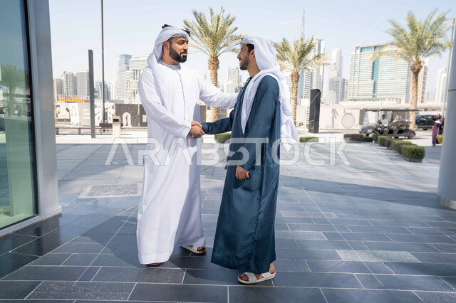 Shaking hands and exchanging conversations between friends, spending quality time with friends, standing confidently on the city streets, enjoying the daytime stroll and breathing in the fresh air, two Emirati Gulf Arab friends wearing white kanduras and ghutras, looking at each other with happy expressions, the concept of elegance and attention to one's outward appearance.