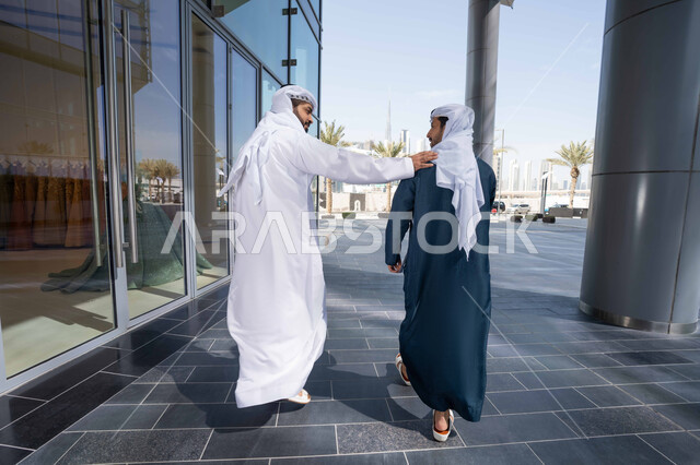 علاقات اجتماعية جديدة ، تبادل الاحاديث بين الاصدقاء ، مفهوم الأناقة والاهتمام بالمظهر الخارجي ، الاستمتاع والتجول واستنشاق الهواء النقي في وقت النهار، الوقوف بثقة في شوارع المدينة، صورة من الخلف لصديقان عربيان خليجيان اماراتيان يرتديان الكندورة والغترة البيضاء ، ينظران الى بعضهما بتعابير السرور .