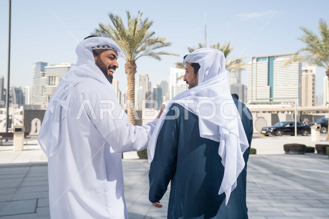 Spending a pleasant time with friends, exchanging conversations between friends, the concept of elegance and taking care of one’s appearance, enjoying, walking around and breathing fresh air during the day, standing confidently in the city streets, a picture from the back of two Emirati Gulf Arab friends wearing the white kandura and ghutra, looking at each other with expressions of pleasure.
