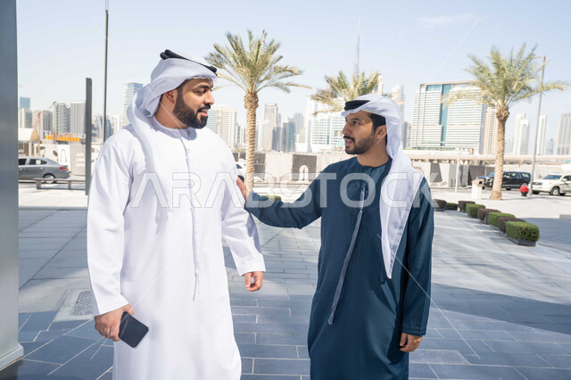 Exchanging conversations between friends, the concept of elegance and interest in one's outward appearance, enjoying, walking around and breathing fresh air during the day, standing confidently in the city streets, spending enjoyable times with friends, two Arab Gulf Emirati friends wearing the white kandura and ghutra, looking at each other with expressions of pleasure.