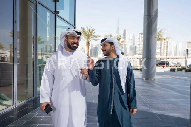 مفهوم الأناقة والاهتمام بالمظهر الخارجي ، الاستمتاع والتجول واستنشاق الهواء النقي في وقت النهار،  الوقوف بثقة في شوارع المدينة، قضاء الاوقات الممتعه مع الاصدقاء ، صديقان عربيان خليجيان اماراتيان يرتديان الكندورة والغترة البيضاء ، ينظران الى بعضهما بتعابير السرور .