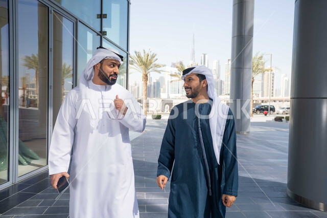 الاستمتاع والتجول واستنشاق الهواء النقي في وقت النهار،  الوقوف بثقة في شوارع المدينة، قضاء الاوقات الممتعه مع الاصدقاء ، صديقان عربيان خليجيان اماراتيان يرتديان الكندورة والغترة البيضاء ، ينظران الى بعضهما بتعابير السرور، مفهوم الأناقة والاهتمام بالمظهر الخارجي .