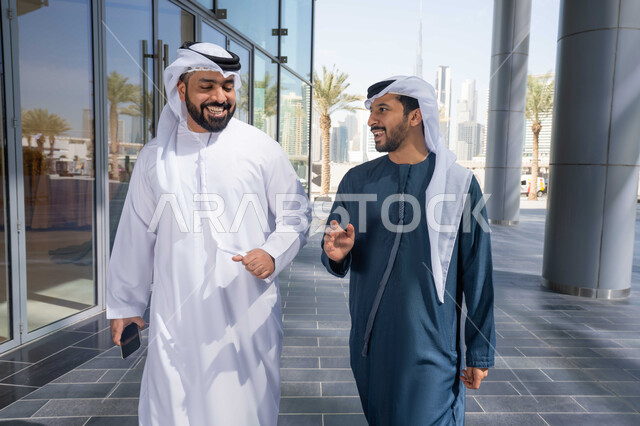Standing confidently in the city streets, spending quality time with friends, enjoying walking around and breathing fresh air during the day, two Emirati Gulf Arab friends wearing white kanduras and ghutras, looking at each other with expressions of pleasure, the concept of elegance and interest in one's outward appearance.