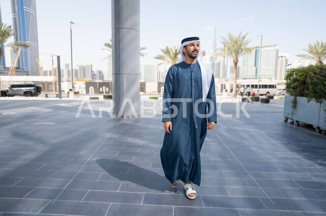 Pride in architectural progress, strolling and breathing fresh air outside, urban growth in the city, and having a good time in the Emirates. An Emirati Gulf Arab man wearing a kandura and white ghutra looks at something during the day.