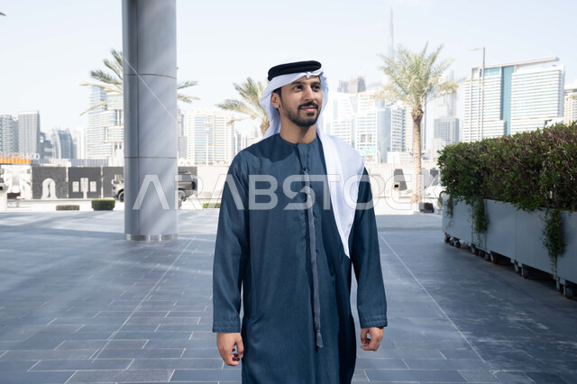 النمو العمراني في المدينة ، قضاء وقت ممتع ف الامارات ، الفخر والاعتزاز بالتقدم المعماري ، التجول واستنشاق الهواء النقي في الخارج,  رجل عربي خليجي اماراتي يرتدي الكندورة والغترة البيضاء ينظر الى شيء ما في وقت النهار .