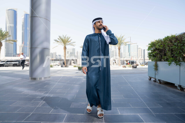 الاطمئنان على الاهل والاصدقاء، استخدام التكنولوجيا والتقنيات الحديثه، نزهة وجولة فى الهواء الطلق ، برامج وتطبيقات الانترنت ، الدردشة مع الاصدقاء ، توثيق اللحظات السعيدة، رجل عربي خليجي اماراتي يرتدي الكندورة والغترة البيضاء، ينظر بتعابير السرور يقوم باستخدام الهاتف المحمول ، التسوق عبر تطبيقات الاونلاين  .