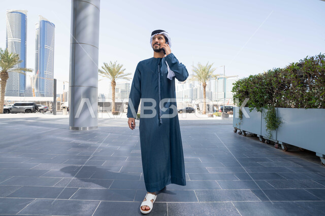 اجراء مكالمة هاتفية ، استخدام التكنولوجيا والتقنيات الحديثه، نزهة وجولة فى الهواء الطلق ، برامج وتطبيقات الانترنت ، الدردشة مع الاصدقاء ، توثيق اللحظات السعيدة، رجل عربي خليجي اماراتي يرتدي الكندورة والغترة البيضاء، ينظر بتعابير السرور يقوم باستخدام الهاتف المحمول ، التسوق عبر تطبيقات الاونلاين  .