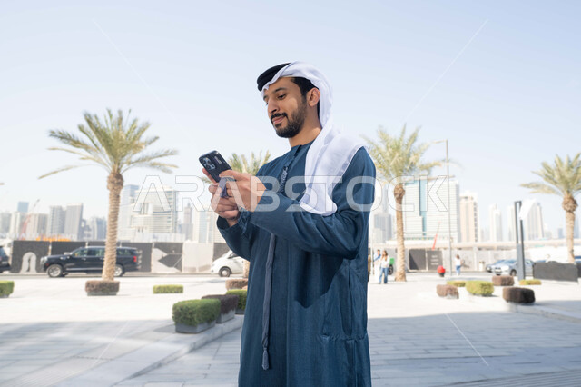 نزهة وجولة فى الهواء الطلق ، برامج وتطبيقات الانترنت ، الدردشة مع الاصدقاء ، توثيق اللحظات السعيدة، استخدام الهاتف المحمول والتقنيات الحديثه، رجل عربي خليجي اماراتي يرتدي الكندورة والغترة البيضاء، ينظر بتعابير السرور يقوم باستخدام الهاتف المحمول ، التسوق عبر تطبيقات الاونلاين  .