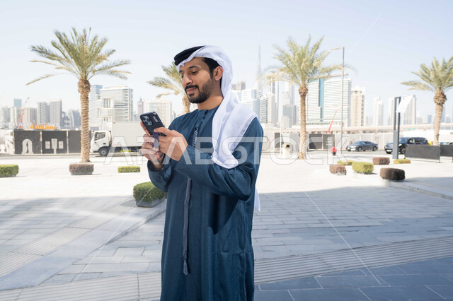 برامج وتطبيقات الانترنت ، الدردشة مع الاصدقاء ، توثيق اللحظات السعيدة، استخدام الهاتف المحمول والتقنيات الحديثه، التسوق عبر تطبيقات الاونلاين ، رجل عربي خليجي اماراتي يرتدي الكندورة والغترة البيضاء، ينظر بتعابير السرور يقوم باستخدام الهاتف المحمول ، نزهة وجولة فى الهواء الطلق .