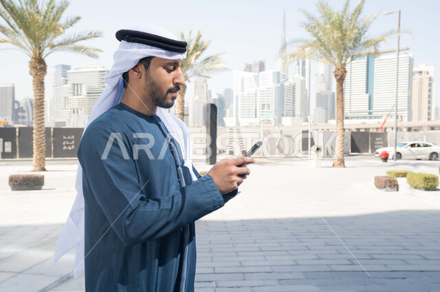 Chatting online, documenting happy moments, using mobile phones and modern technologies, shopping via online applications, an Emirati Gulf Arab man wearing a white kandura and ghutra, looking happy while using his mobile phone, taking a walk and a tour in the open air.