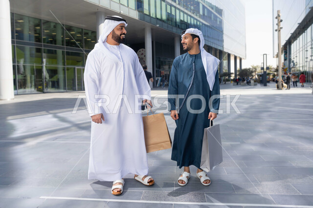 علاقات إجتماعية بين الإصدقاء ، الشراء فى ايام التخفيضات ، ايماءات وتعابير السعادة ، قضاء أوقات ممتعة في الطبيعة ، التجول واستنشاق الهواء النقي في وقت النهار، صديقان عربيان خليجيان اماراتيان يرتديان الكندورة والغترة البيضاء ،  يحملان اكياس التسوق بتعابير السرور.