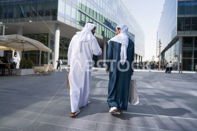 شراء الهدايا ، متعة التسوق ، ايماءات وتعابير السعادة ، قضاء أوقات ممتعة في الطبيعة ، التجول واستنشاق الهواء النقي في وقت النهار، صورة من الخلف لصديقان عربيان خليجيان اماراتيان يرتديان الكندورة والغترة البيضاء ،  يحملان اكياس التسوق بتعابير السرور.