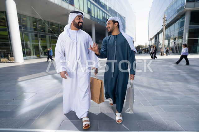 The joy of shopping with friends, spending quality time in nature, walking around and breathing fresh air during the day, making friends, getting to know each other, discussions and dialogues, the concept of elegance and taking care of one's appearance, two Arab Gulf Emirati friends wearing the white kandura and ghutra, carrying shopping bags with happy expressions.
