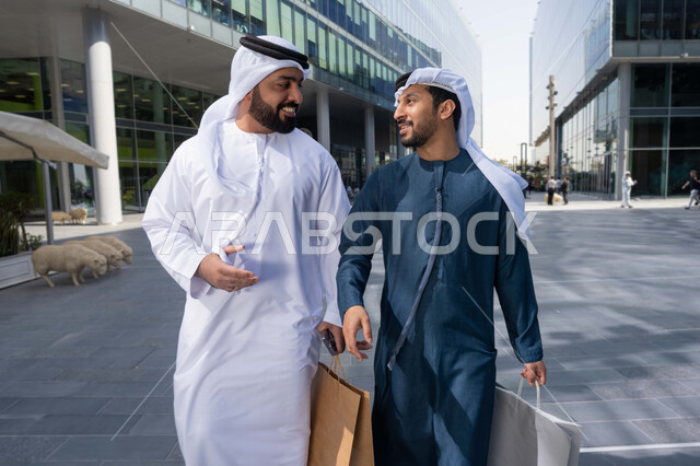 الاستمتاع والتجول واستنشاق الهواء النقي في وقت النهار، صداقات وتعارف ونقاشات وحوارات ، قضاء أوقات ممتعة في الطبيعة ، مفهوم الأناقة والاهتمام بالمظهر الخارجي ،  صديقان عربيان خليجيان اماراتيان يرتديان الكندورة والغترة البيضاء ،  يحملان اكياس التسوق بتعابير السرور.