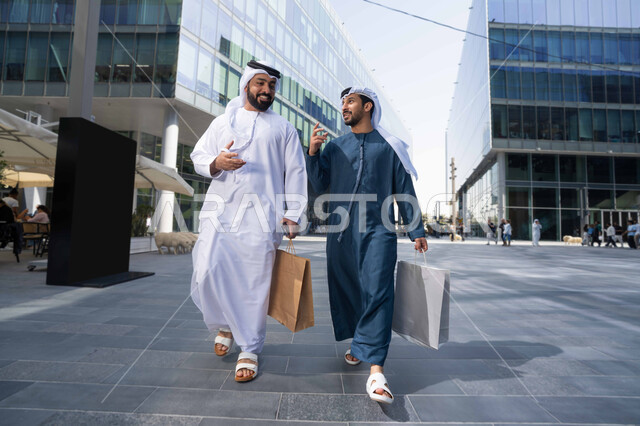 صداقات وتعارف ونقاشات وحوارات قضاء أوقات ممتعة في الطبيعة ، مفهوم الأناقة والاهتمام بالمظهر الخارجي ، الاستمتاع والتجول واستنشاق الهواء النقي في وقت النهار، صديقان عربيان خليجيان اماراتيان يرتديان الكندورة والغترة البيضاء ،  يحملان اكياس التسوق بتعابير السرور.