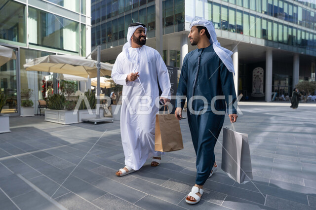 مفهوم الأناقة والاهتمام بالمظهر الخارجي ، صداقات وتعارف ونقاشات وحوارات قضاء أوقات ممتعة في الطبيعة ، الاستمتاع والتجول واستنشاق الهواء النقي في وقت النهار، صديقان عربيان خليجيان اماراتيان يرتديان الكندورة والغترة البيضاء ،  يحملان اكياس التسوق بتعابير السرور.