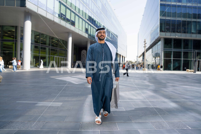 النمو والتطور العمراني في الامارات ، الفخر والاعتزاز بالتقدم المعماري ، التجول واستنشاق الهواء النقي في الخارج, صورة لرجل عربي خليجي اماراتي يرتدي الكندورة والغترة البيضاء ينظر الى شيء ما في وقت النهار ، يحمل اكياس التسوق .