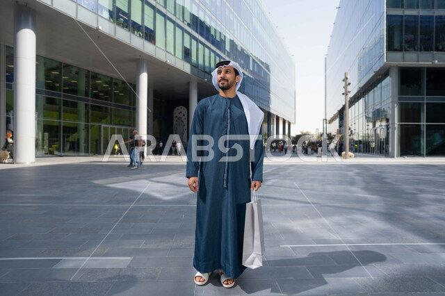 Watching the urban development in the city, urban growth in the Emirates, pride and admiration for the architectural progress, walking around and breathing the fresh air outside, a picture of an Emirati Gulf Arab man wearing a white kandura and ghutra looking at something during the day, carrying shopping bags.