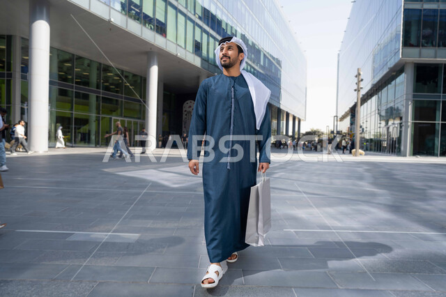 الفخر والاعتزاز بالتقدم المعماري ، التجول واستنشاق الهواء النقي في الخارج, النمو والتطور العمراني في الامارات ، صورة لرجل عربي خليجي اماراتي يرتدي الكندورة والغترة البيضاء ينظر الى شيء ما في وقت النهار ، يحمل اكياس التسوق .