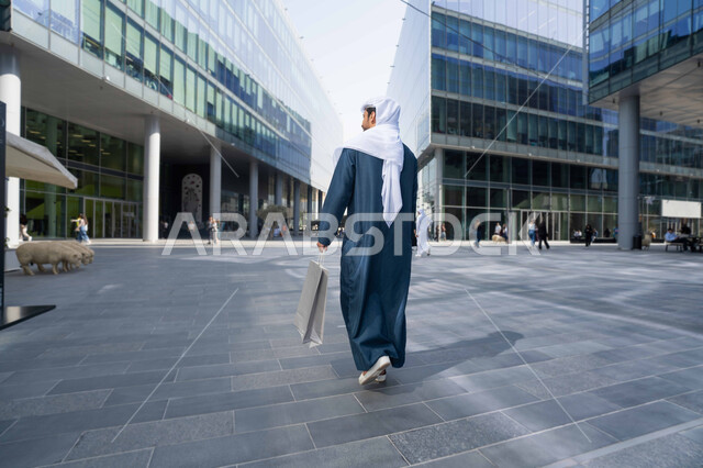 قضاء وقت ممتع ف الامارات ، النمو العمراني في الامارات ، الفخر والاعتزاز بالتقدم المعماري ، التجول واستنشاق الهواء النقي في الخارج, صورة من الخلف لرجل عربي خليجي اماراتي يرتدي الكندورة والغترة البيضاء ينظر الى شيء ما في وقت النهار ، يحمل اكياس التسوق .