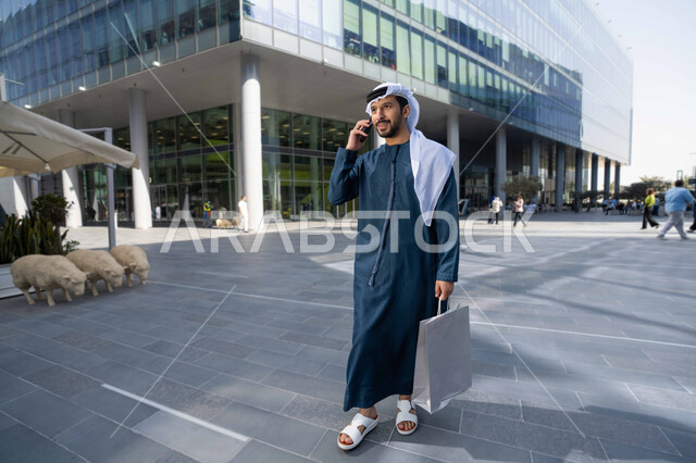 ايماءات السعادة لسماع الاخبار المبهجة  ، برامج وتطبيقات الجوال المتنوعة ، اجراء مكالمة هاتفية عبر الجوال ، الاطمئنان على الأصدقاء، تبادل الاحاديث والاخبار بين العائلة، رجل عربي خليجي اماراتي يقوم بإستخدام الأجهزة التقنية الحديثة، يحمل اكياس التسوق .
