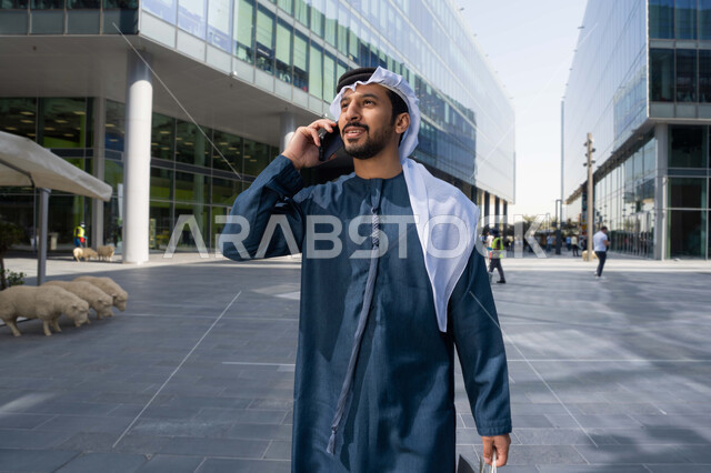 Various mobile programs and applications, making a phone call via mobile, checking on friends, exchanging conversations and news with family, an Arab Gulf Emirati man using modern technical devices, carrying shopping bags.